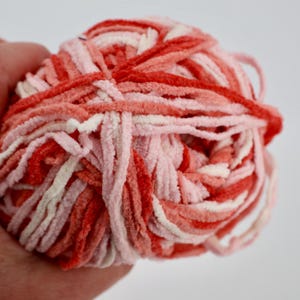 May include: A ball of soft, plush chenille yarn featuring a variegated color palette of bright red, coral, light pink, and white strands twisted together. The texture is fuzzy and velvety, creating a thick, rounded bundle of craft material.