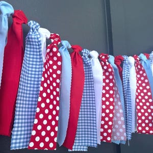 May include: A festive fabric banner with alternating red and white polka dot, solid red, light blue, and blue gingham fabric strips. The banner is tied at the top with knots, creating a decorative garland.
