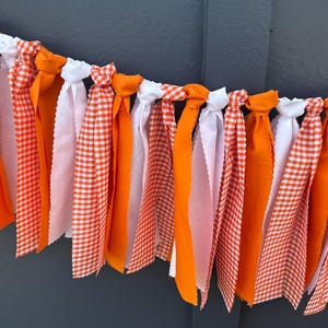 Może przedstawiać: Dekoracyjny baner z naprzemiennymi pomarańczowymi, białymi oraz pomarańczowo-białymi paskami materiału w kratę. Paski materiału są zawiązane na białym sznurku, tworząc świąteczną i kolorową girlandę. Idealna na imprezy.