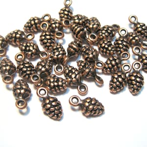 May include: A group of 25 copper-colored metal pine cone charms with a loop for hanging. The charms are about 1 inch long and have a detailed, textured surface.