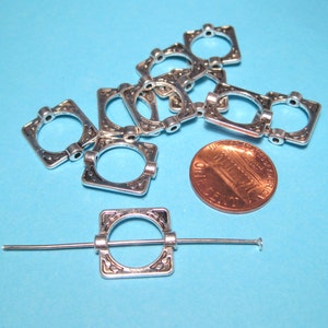 May include: A close-up of silver-toned metal square-shaped jump rings with ornate designs. The jump rings are arranged in a pile on a blue surface. A penny is included for size reference.