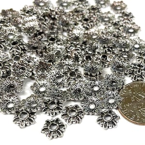 Puede incluir: Un mont&oacute;n de tapas de cuentas de metal plateado con un dise&ntilde;o floral decorativo. Las tapas de cuentas son aproximadamente del tama&ntilde;o de una moneda de euro.