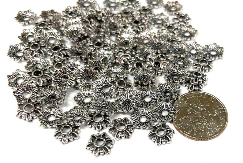 Puede incluir: Un mont&oacute;n de tapas de cuentas de metal plateado con un dise&ntilde;o floral decorativo. Las tapas de cuentas son aproximadamente del tama&ntilde;o de una moneda de euro.
