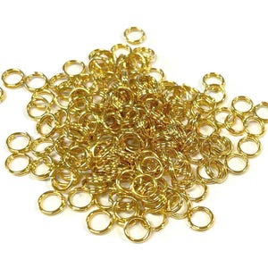 May include: A collection of small, gold-coloured split rings, suitable for jewellery making or crafts. The rings are tightly closed and have a bright, shiny finish. They are scattered on a white background, creating a visually appealing arrangement.