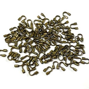 May include: A pile of small, antique brass-colored metal clasps. Each clasp has a U-shaped design with a small, cylindrical barrel at each end. The clasps are scattered on a white surface, creating a textured appearance.