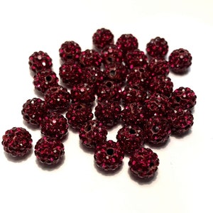 May include: A collection of burgundy-colored, spherical beads. Each bead is covered in small, sparkling crystals, giving them a textured appearance. The beads are scattered on a white surface, creating a visually appealing display.