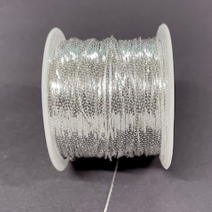 May include: A spool of silver chain, wound around a clear plastic bobbin. The chain is made of small, flat, rectangular links.