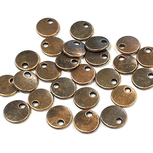 May include: A collection of small, round, bronze-colored metal charms with a hole at the top for stringing. The charms have a slightly aged appearance, with variations in color and texture. They are scattered on a white surface.