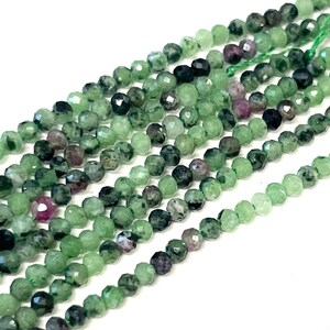 May include: Strands of faceted gemstone beads in shades of green, dark green, and hints of purple. The beads are strung on a green thread, creating a textured and colorful display. Ideal for jewelry making.