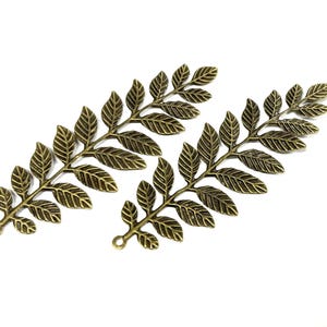 Pode incluir: Três pingentes de folhas de metal cor bronze com veios detalhados. Cada pingente tem um pequeno laço na base para prender a joias ou outros projetos de artesanato. As folhas estão dispostas sobre um fundo branco.