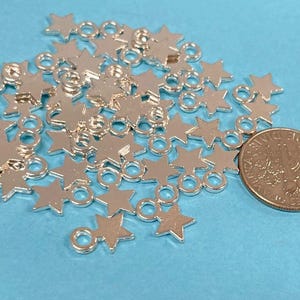 May include: A pile of silver-toned metal star charms with jump rings. The charms are scattered on a light blue surface. A US quarter is included for size reference.