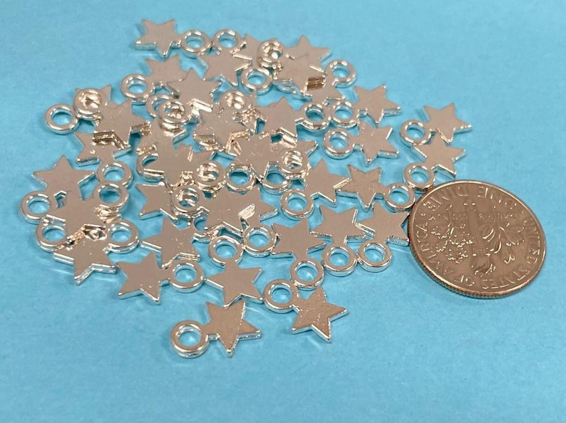 May include: A pile of silver-toned metal star charms with jump rings. The charms are scattered on a light blue surface. A US quarter is included for size reference.