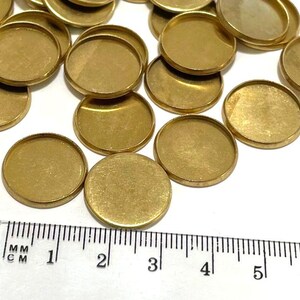 Puede incluir: Primer plano de un grupo de 20 engastes redondos de metal de color dorado. Los engastes tienen aproximadamente 2,5 cm de di&aacute;metro y un acabado liso y pulido. Se incluye una regla para la escala.