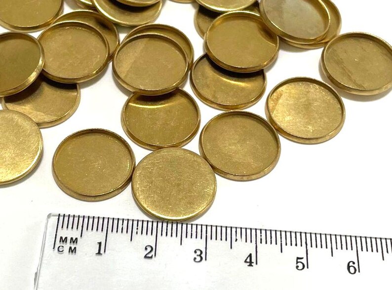 Puede incluir: Primer plano de un grupo de 20 engastes redondos de metal de color dorado. Los engastes tienen aproximadamente 2,5 cm de di&aacute;metro y un acabado liso y pulido. Se incluye una regla para la escala.