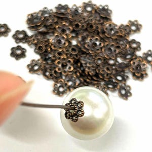 May include: A close-up of a white pearl bead with a bronze flower-shaped bead cap. A pile of bronze flower-shaped bead caps are scattered in the background.