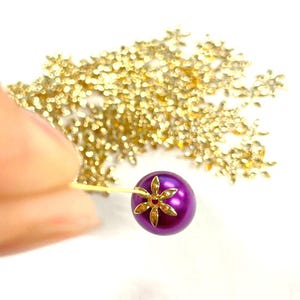 以下が含まれることがあります： 中央に金のフラワーデザインが施された紫色の真珠ビーズ1つ。ビーズは金色のワイヤーに付いています。