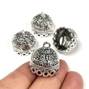 May include: Silver-colored metal bell-shaped beads with intricate floral and geometric patterns. The beads have a small loop at the top and a series of circular cutouts around the base. One bead is held in a hand.