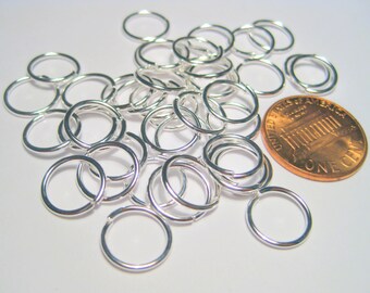 May include: A pile of silver jump rings, some overlapping, next to a US penny. The jump rings are small, about the size of a penny.