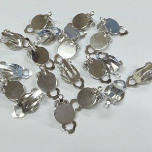 May include: A collection of silver-colored clip-on earring findings. Each finding has a round, flat base with a loop and a hinged clip mechanism. The findings are scattered across a white surface, reflecting light.