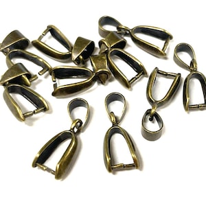May include: A collection of antique brass-colored pendant bails, ideal for jewelry making. The bails have a loop at the top and a hinged clasp at the bottom. The bails are scattered on a white surface.