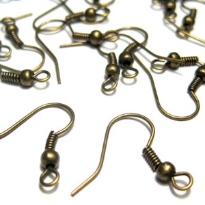 May include: Close-up of several antique bronze-colored earring hooks. The hooks have a curved design with a coiled spring and a small ball at the end. The hooks are scattered on a white surface, showcasing their intricate details.