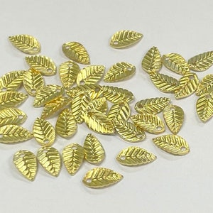 May include: A collection of gold-coloured metal leaf charms with a hole in the centre. The charms are scattered on a white surface.