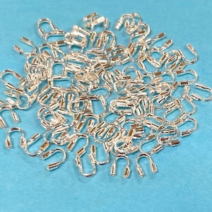May include: A large pile of silver-toned metal crimp beads. The beads are small and have a loop on one end and a closed end on the other.