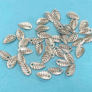 May include: A pile of silver metal leaf charms with a hole in the center of each leaf. The leaves are arranged in a random pattern on a light blue background.