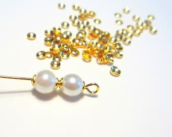 May include: Two white pearl beads strung on a gold wire with a gold loop at the end. A pile of gold jump rings are scattered on a white surface.