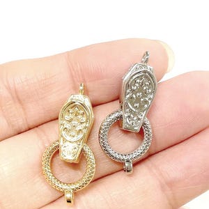May include: Two ornate metal jewelry clasps. One is gold-toned, the other silver-toned. Each clasp features a decorative coffin-shaped top with an intricate design and a textured circular ring. The clasps are held in a hand against a white background.