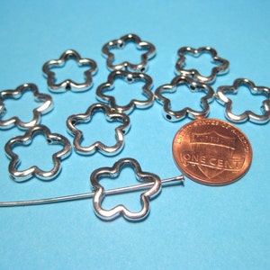 May include: A group of ten silver metal flower-shaped charms, each with a hole in the center, are scattered on a blue surface. A penny is included for size comparison.