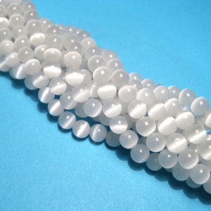 May include: A strand of white round beads with a smooth, glossy finish. The beads have a subtle shimmer and are arranged in a loose, flowing pattern.