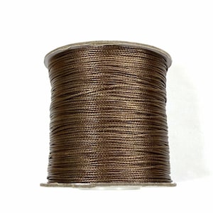 Pode incluir: Um carretel de cordão castanho, bem enrolado. O cordão parece ser feito de um material sintético liso, enrolado em torno de um carretel de cartão. A cor é um castanho quente e rico, adequado para trabalhos manuais.