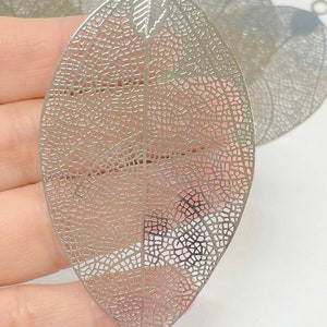 以下が含まれることがあります： 繊細で複雑なデザインの銀色の金属製の葉のチャーム。葉の上部に吊り下げ用の穴があります。