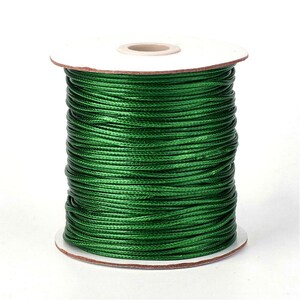 May include: A spool of green waxed cotton cord. The cord is wound tightly around a white cardboard spool.