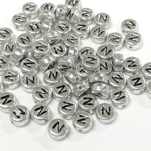 May include: Silver-colored, round beads with the letter "N" in black. The beads are scattered, showing a metallic finish and a small hole for stringing. These beads are suitable for jewelry making and crafts.