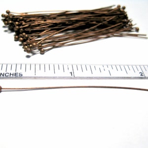 Puede incluir: Un mont&oacute;n de 100 alfileres de cabeza dorada, cada uno de 2,5 cm de largo. Los alfileres est&aacute;n dispuestos en un mont&oacute;n suelto sobre una superficie blanca. Una regla se coloca debajo del mont&oacute;n de alfileres para mostrar el tama&ntilde;o de los alfileres.