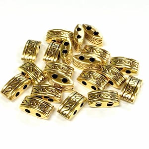 May include: A collection of gold-coloured metal beads with an ornate, swirling design. Each bead has two holes for stringing and a rectangular or oval shape. The beads are scattered on a white surface.