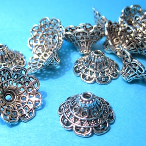May include: A group of ten silver filigree metal bead caps with a decorative design. The caps are arranged on a blue background.