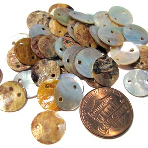 May include: A collection of iridescent, round shell beads with two holes each. The beads vary in color, including shades of brown, gold, and pearlescent white. A US one-cent coin is included for size reference.