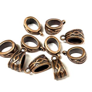 May include: Ten copper-toned metal pendant bails with an intricate design. The bails are oval-shaped with a loop at the top for attaching a pendant.