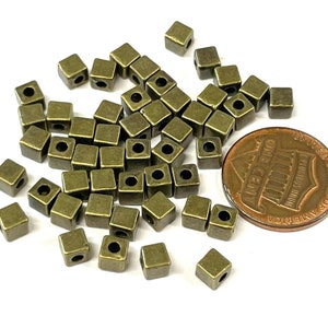 May include: A pile of antique gold metal square beads with a hole in the center of each bead. The beads are scattered on a white surface next to a US penny.