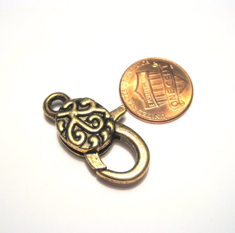 May include: A gold-toned metal clasp with a decorative swirl design. The clasp is about the size of a US penny.
