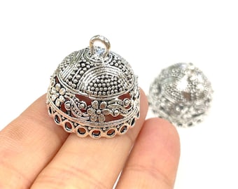 May include: Silver-colored, dome-shaped jewelry component with intricate floral and dot patterns. The component has a small loop at the top and a decorative edge. Another similar component is visible in the background.
