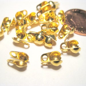 May include: A close-up of a group of gold-toned metal bead caps with a small loop on each side. The bead caps are scattered on a white surface.