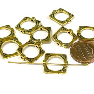 May include: A collection of gold-colored square metal jewelry findings with decorative black accents. The findings have a square shape with a circular opening in the center. A penny is included for size reference. A gold pin is also included.
