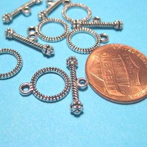 Op de afbeelding: Zilverkleurige metalen toggle-sluitingen met een touwontwerp. De sluitingen zijn ongeveer zo groot als een Nederlandse cent.