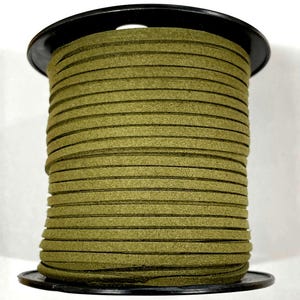 May include: A spool of olive green suede leather cord. The cord is tightly wound around a black plastic spool. The suede appears to be flat and uniform in width. The cord is suitable for jewelry making, crafting, or other creative projects.