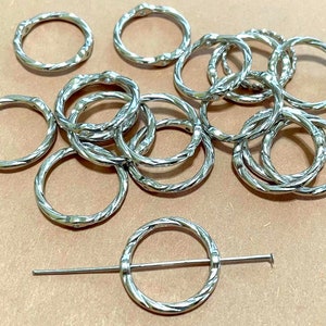 May include: A collection of silver-colored metal shawl pins and rings. The rings have a textured, twisted design. One pin is inserted through a ring. The items are displayed on a light brown surface.
