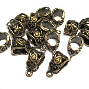 May include: A group of 12 antique gold-toned metal pendant bails with ornate designs. The bails have a loop at the top and a small opening at the bottom for attaching a pendant.